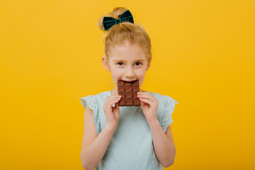 little girl tasting chocolate, look at the camera, in blue T-shirt, isolated yellow background, copy space