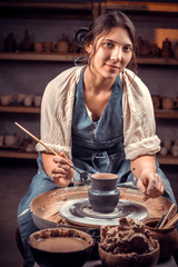 Wall Mural - Stylish young and cheerful woman working on potter's wheel with raw clay with hands. Making ceramic dishes.