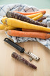 Bunte Urkarotte und Möhren in Lila, Orange und Gelb auf Geschirrtuch mit Schäler auf Holzbrett 