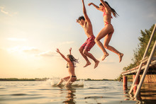 A Group Of Young People Joyfully Leap Into The Refreshing Lake Waters On A Sunny Summer Day, Embracing The Carefree Moments Of Pure Fun And Laughter.