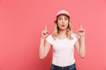 Poster - Portrait of stylish blond woman 20s wearing casual t-shirt and cap pointing fingers upward at copyspace isolated over pink background