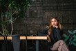  A sophisticated, young, tanned professional Southeast Asian business woman in a turtle neck top looking at the viewer and is sitting on a bench in the city restaurant during the day.
