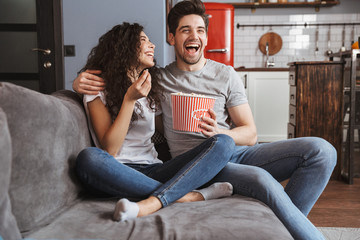 Wall Mural - Picture of joyful young couple sitting on sofa at home and eating popcorn from bucket