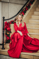 Wall Mural - christmas portrait of beautiful woman with blond hair, wearing long red dress, sitting on stairs