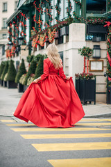Wall Mural - christmas portrait of beautiful woman with blond hair, wearing long red dress, walking down the stre