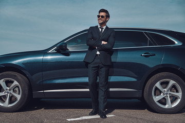 Wall Mural - Confident in his style. Full length of handsome young businessman keeping arms crossed and smiling while standing near his car outdoors