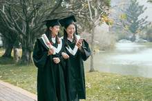 Happy Graduates. Two Asian Beautiful Women In Graduation Gowns Looking At Mobile Phone Together Smiling Standing On Little Path Way By Pond Outdoor On Sunny Day. Girls Sharing Cellphone Search Job.