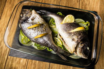 two Dorada fish cooked in the oven with lemon, onion and green pimento in a glass dish on a black slate tray