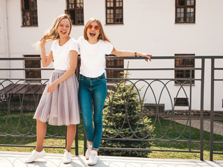 Portrait of two young beautiful blond smiling hipster girls in trendy summer white t-shirt clothes. Sexy carefree women posing on street background. Positive models having fun in sunglasses.Hugging