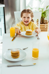 Wall Mural - selective focus of happy excited kid sitting at table near orange juice and holding plate with pancakes