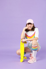 Wall Mural - Smiling teen girl in vivid clothes sitting holding yellow skateboard put hand prop up on chin isolated on violet pastel wall background. People sincere emotions, lifestyle concept. Mock up copy space.