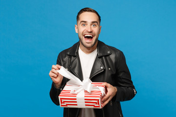 Handsome stylish young bearded man in black leather jacket white t-shirt hold gift box isolated on blue wall background studio portrait. People sincere emotions lifestyle concept. Mock up copy space.