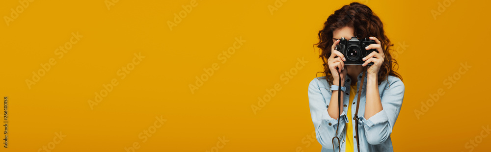 panoramic shot of curly redhead photographer covering face with digital camera isolated on orange - obrazy, fototapety, plakaty 