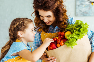 Wall Mural - happy mother showing smiling daughter paper bag with ripe fruits and vegetables