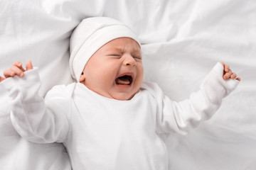 screaming baby in white clothes lying on bed with closed eyes