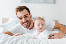 Laughing Dad Lying On Bed With Cute Baby And Looking At Camera