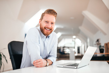 Wall Mural - Handsome millennial redhead man smiling at camera while sitting at desk with laptop.