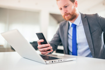 Wall Mural - Redhead businessman using smart phone and laptop at work, close-up.