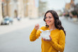 © bnenin - Millennial woman eating takeout food in the city street, smiling at camera.