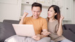 © tirachard - Asian couple using laptop for search web in living room at home, sweet couple enjoy love moment while lying on the sofa when relax at home. Lifestyle couple relax at home concept.