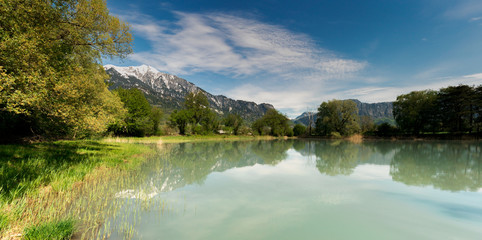 Canvas Print - springtime landscape with snowcapped mountains and pond in green forest