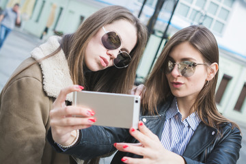 two stylish girls take a selfie on their smartphone on a city street and smile at the camera