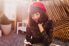Adorable Smiling Girl With Brown Short Hair Sitting At Outdoor Cafe And Texting Message In Sunny Day. Portrait Of Cheerful Young Brunette Woman In French Vintage Outfit Resting At Open-air Restaurant