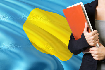 Wall Mural - Learning Palauan language concept. Young woman standing with the Palau flag in the background. Teacher holding books, orange blank book cover.