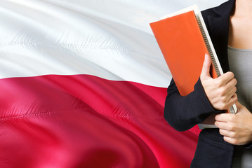 Wall Mural - Learning Polish language concept. Young woman standing with the Poland flag in the background. Teacher holding books, orange blank book cover.