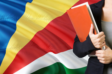 Wall Mural - Learning Seychellois language concept. Young woman standing with the Seychelles flag in the background. Teacher holding books, orange blank book cover.