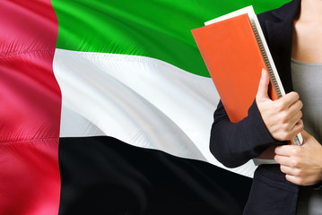 Wall Mural - Learning Emirian language concept. Young woman standing with the United Arab Emirates flag in the background. Teacher holding books, orange blank book cover.