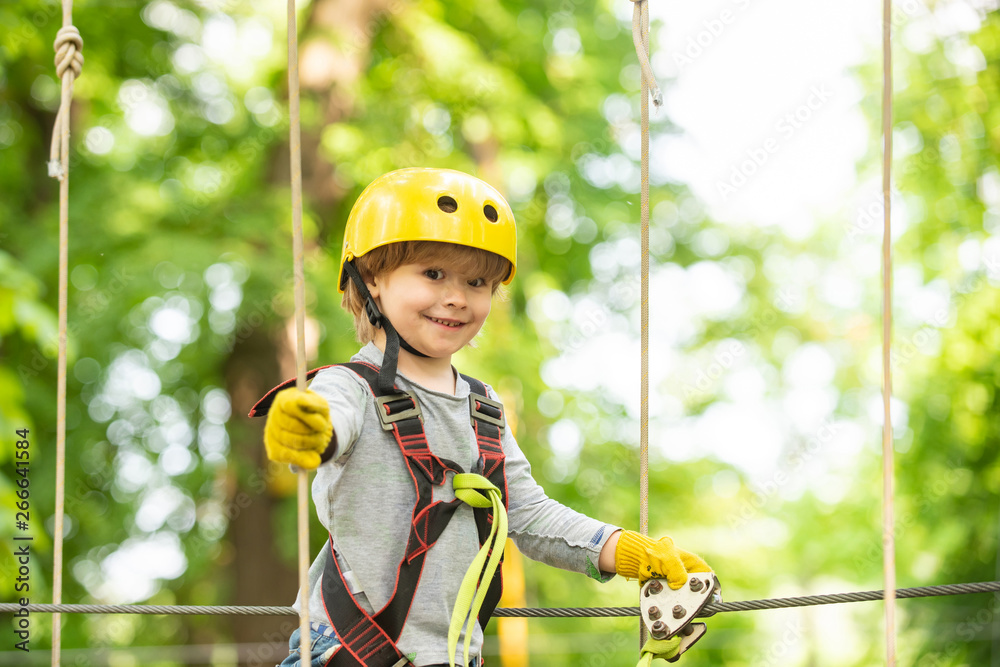 Happy child boy calling while climbing high tree and ropes. Portrait of a beautiful kid on a rope park among trees. Small boy enjoy childhood years. - obrazy, fototapety, plakaty 