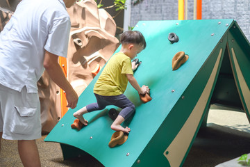 asian father & cute 2 - 3 years old toddler child having fun trying to climb on artificial boulders 