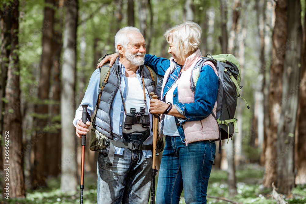Lovely senior couple hugging in the forest while hiking with backpacks and trekking sticks. Concept of active lifestyle on retirement - obrazy, fototapety, plakaty 