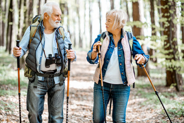 Wall Mural - Beautiful senior couple hiking with backpacks and trekking sticks in the forest. Concept of an active lifestyle on retirement