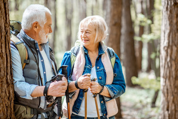 Wall Mural - Senior couple resting near the tree while traveling with backpacks and trekking sticks in the forest. Concept of an active lifestyle on retirement