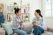 © PR Image Factory - two happy young asian female friends with coffee cups conversing in living room at home. group of beautiful girls bestie sitting on couch relax in bright cozy apartment enjoy tea in weekends gossip