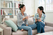 group of happy asian female friends talking and laughing loud holding drinks sitting on sofa in the living room at home. young girls sisters gossip having fun on couch with tea cups indoors apartment