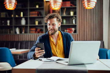 Young Caucasian businessman dressed smart casual using smart phone while sitting in cafe. On the desk laptop.