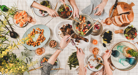 Wall Mural - Family or friends gathering dinner. Flat-lay of peoples hands, roasted lamb shoulder, salads, vegetables, rose wine, mimosa branches over white checkered tablecloth, top view. Celebration party dinner