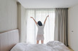 © anon - Woman stretching in bed after waking up, back view. Woman sitting near the big white window while stretching on bed after waking up with sunrise at morning, back view.
