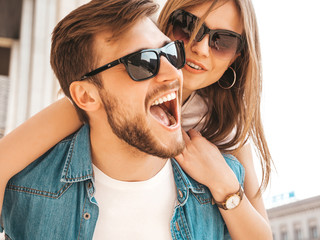 Smiling beautiful girl and her handsome boyfriend in casual summer clothes. Man carrying his girlfriend on the back and she raising her hands.Happy cheerful family having fun on the street background