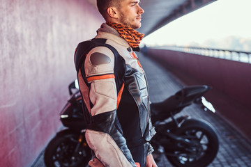 Young pensive biker is standing near his motobike in quiet tunnel. He is wearing special gear and scarf.