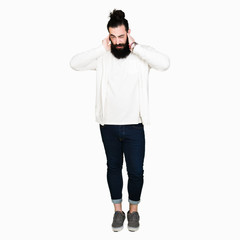 Wall Mural - Young man with long hair and beard wearing sporty sweatshirt covering ears with fingers with annoyed expression for the noise of loud music. Deaf concept.