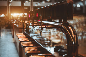 True tilt-shift view of multiple brassy beer taps for lager beer, and craft beer in an indoors restaurant with blank black screens on the top of each tap, thick gilded pipe and glasses behind it