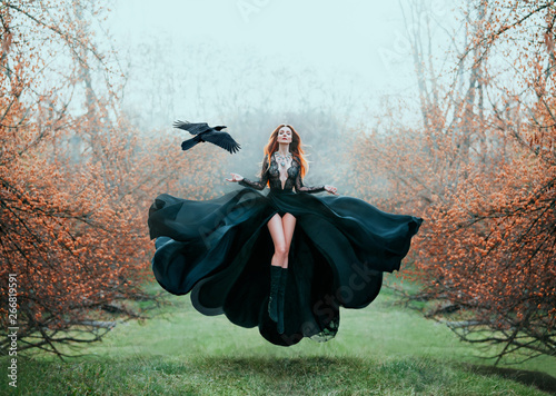girl with bright red hair levitates above ground, powerful sorceress, forest goddess in black flying dress with lace on open chest and bare long legs, lady in flowering orange garden and hand raven
