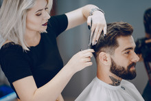 Handsome Young Bearded Guy Sitting In An Armchair In A Beauty Salon And The Girl Near Him Cuts His Hair