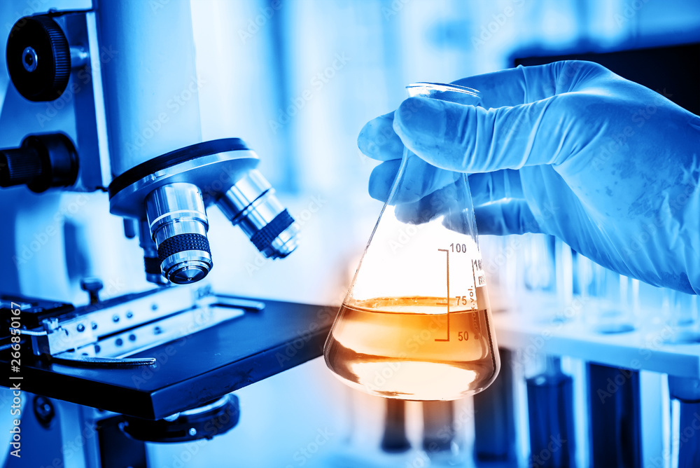 Flask in scientist hand with lab microscope background in laboratory. Science or chemical research and development concept.