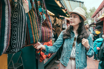 Wall Mural - Summer holidays shopping concept. Young asian woman tourist buying souvenirs in gift olvera street stall outdoor picking choosing bags at vendor. girl traveler shopping in mexico market on sunny day