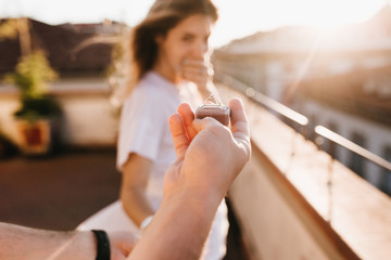 Man holding wedding ring in front of astonished happy girl covering mouth with hand. Romantic photo of charming woman standing on roof early in evening on date with boyfriend in anniversary.